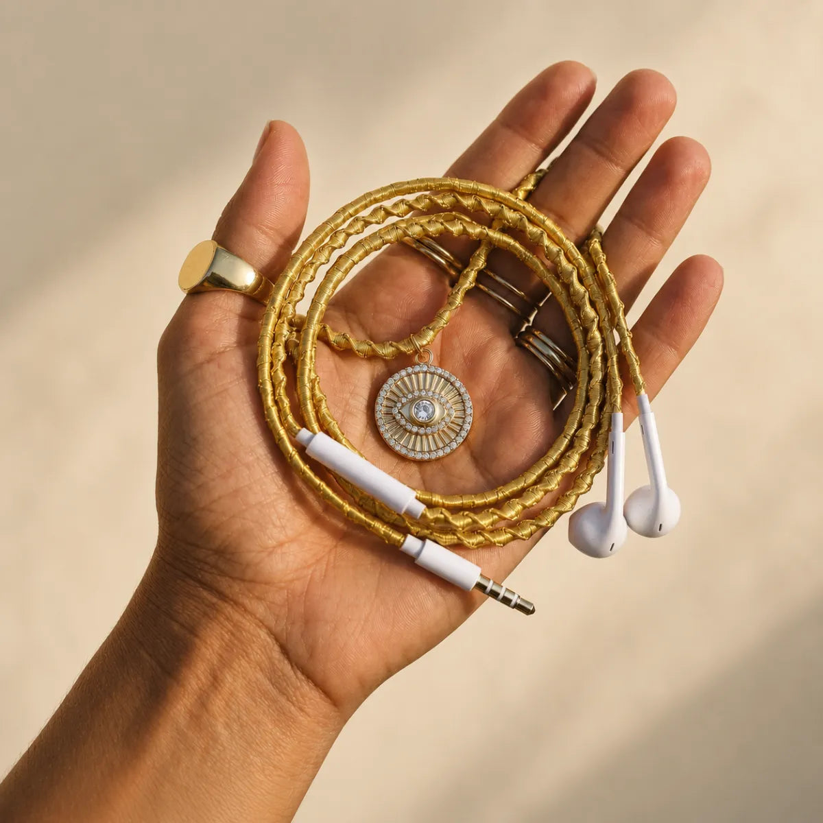 Sun-tanned hand with thin gold stacking rings and chunky signet ring holding gold evil-eye-medallion earphones against soft cream stucco wall