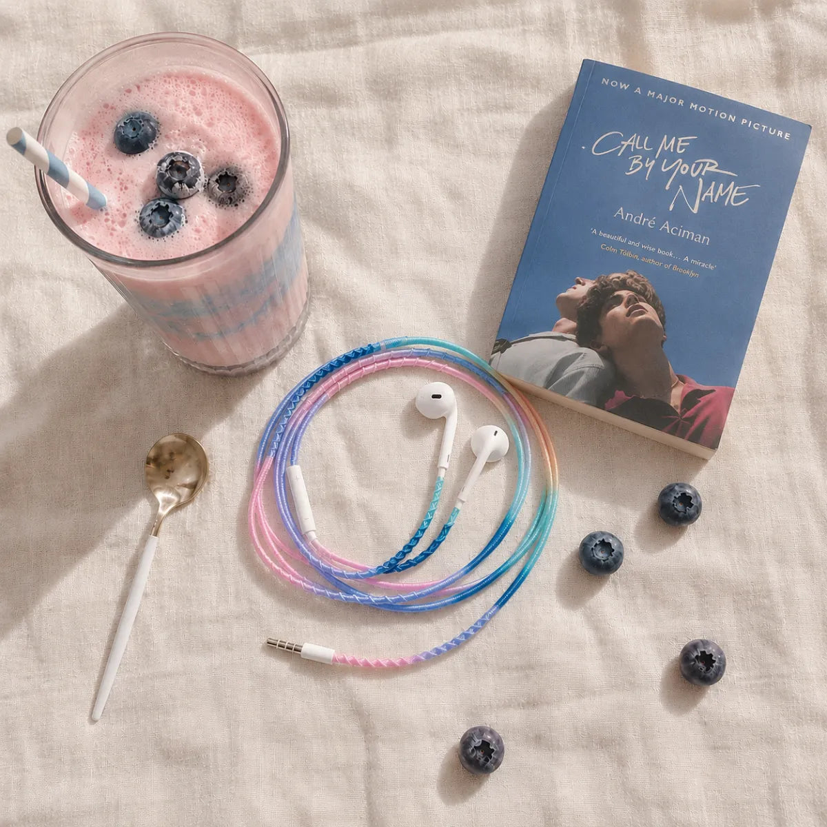 Top-down flat lay of blueberry blush milky gradient wrapped earphones beside a pink and blue milkshake, white hardcover book, and scattered fresh blueberries on cream linen 