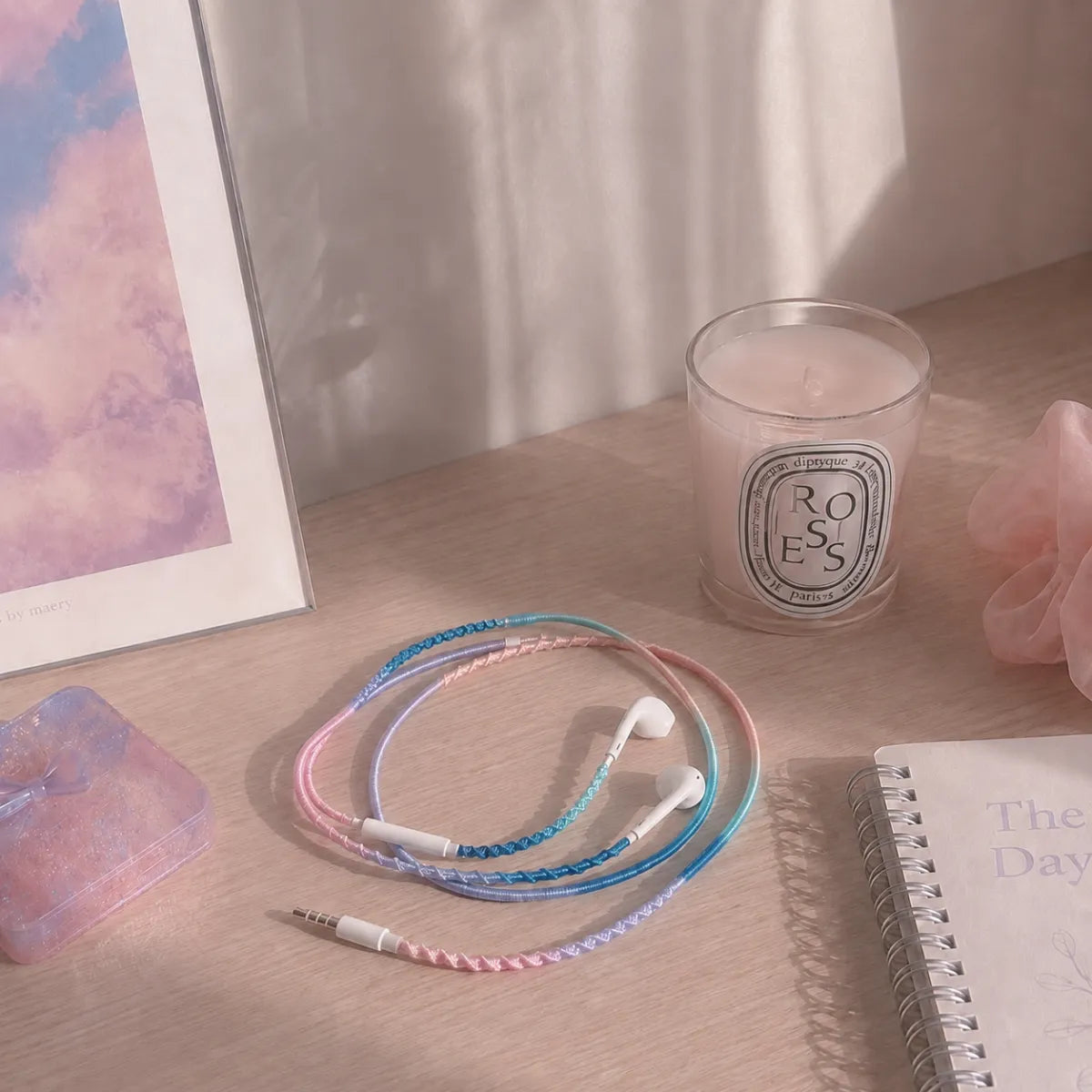  Pale desk corner with blueberry blush milky gradient wrapped earphones coiled at the base of a small white vase and a soft pastel candle in dreamy diffused window light 