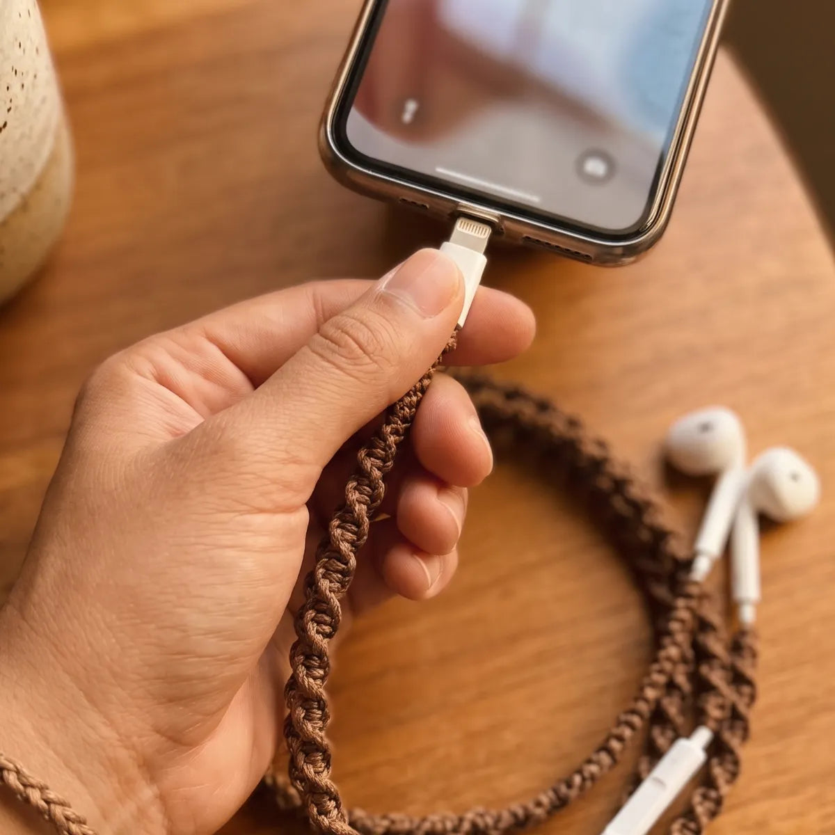  First person POV of a hand plugging natural brown macrame open-knot wrapped earphones into smartphone in warm window light highlighting the chunky handmade knot texture 