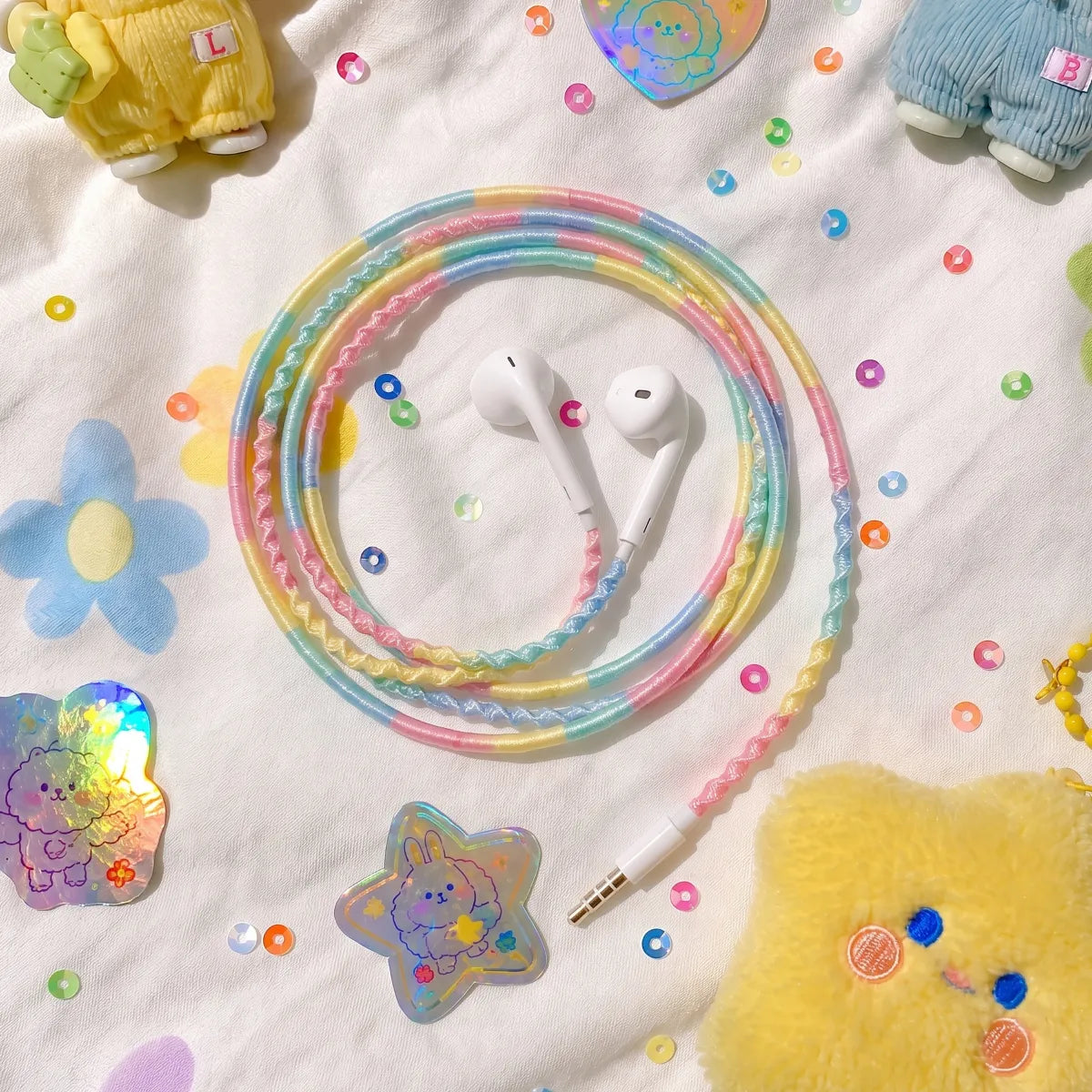  Pastel bedding photo dump with cotton candy four-color block earphones scattered alongside Smiski-style designer toys, sticker sheets, and rainbow sequin scattered across white sheets 