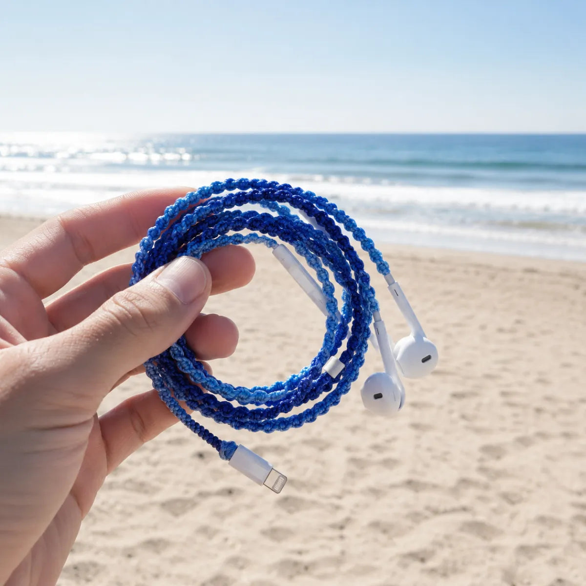  Hand holding deep ocean blue crochet wrapped earphones with white Lightning connector on a sunlit sandy beach with blurred ocean horizon, coastal aesthetic candid 