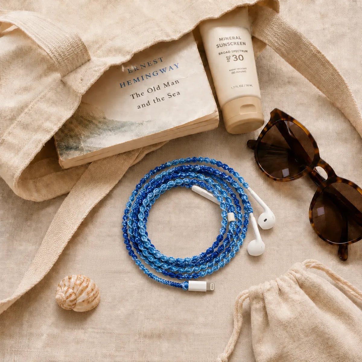  Canvas tote bag contents flat lay with deep ocean blue crochet wrapped earphones beside sunscreen tube, tortoise sunglasses, paperback book, and a small seashell on sand-toned linen 