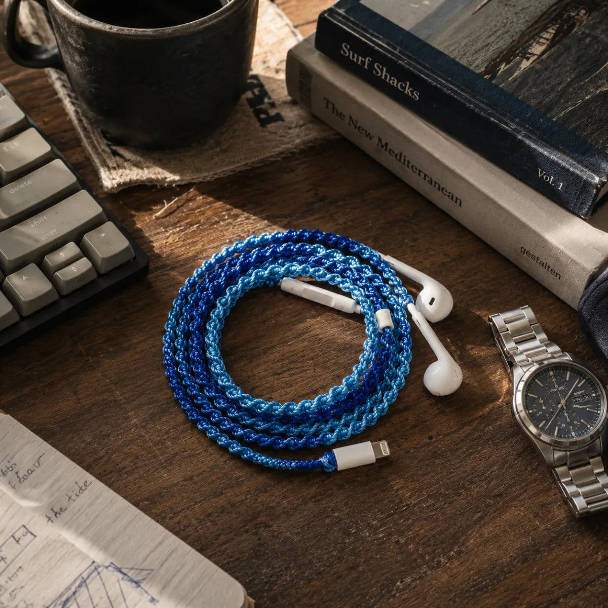  Deep ocean blue crochet wrapped earphones coiled beside a dark mechanical keyboard, ceramic mug of black coffee, and a stack of books on a moody wood desk in window light 