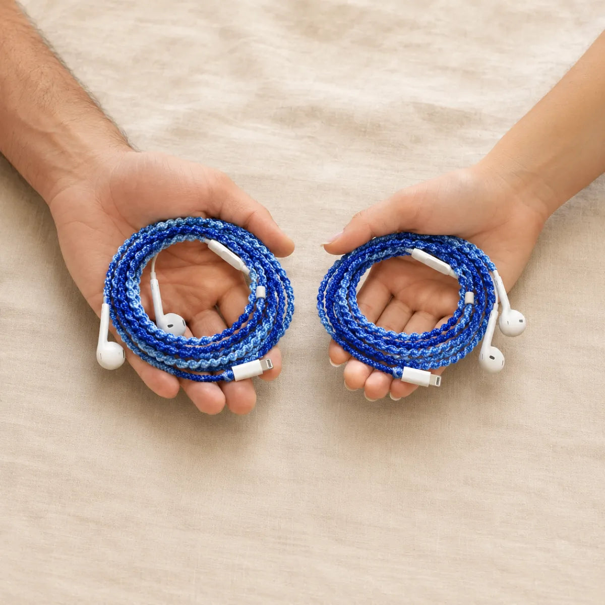  Two hands one male one female each holding identical deep ocean blue crochet wrapped earphones against cream linen background showing unisex coastal style 