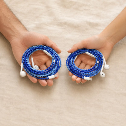  Two hands one male one female each holding identical deep ocean blue crochet wrapped earphones against cream linen background showing unisex coastal style 