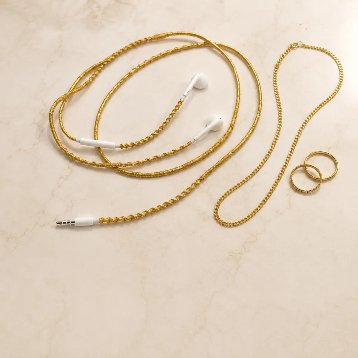  Gold wrapped earphones coiled beside fine gold jewelry including chain necklaces and stacking rings on warm marble surface for quiet luxury aesthetic 
