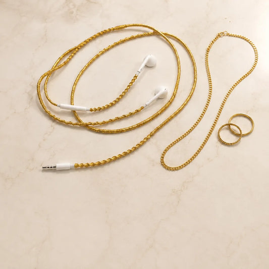  Gold wrapped earphones coiled beside fine gold jewelry including chain necklaces and stacking rings on warm marble surface for quiet luxury aesthetic 