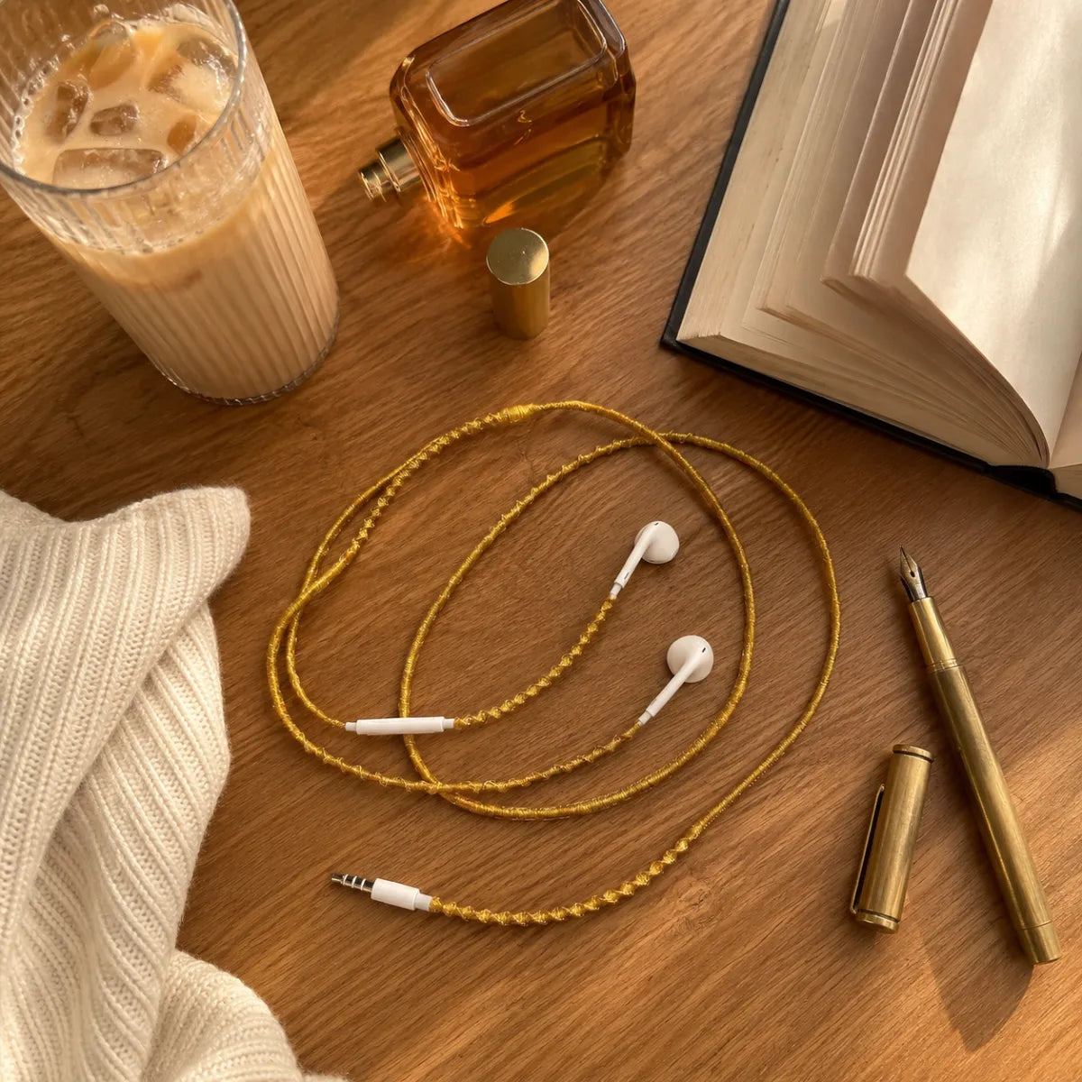  Gold wrapped earphones coiled on warm wooden desk beside iced latte glass, brass pen, and uncapped perfume bottle in clean girl workspace flat lay 