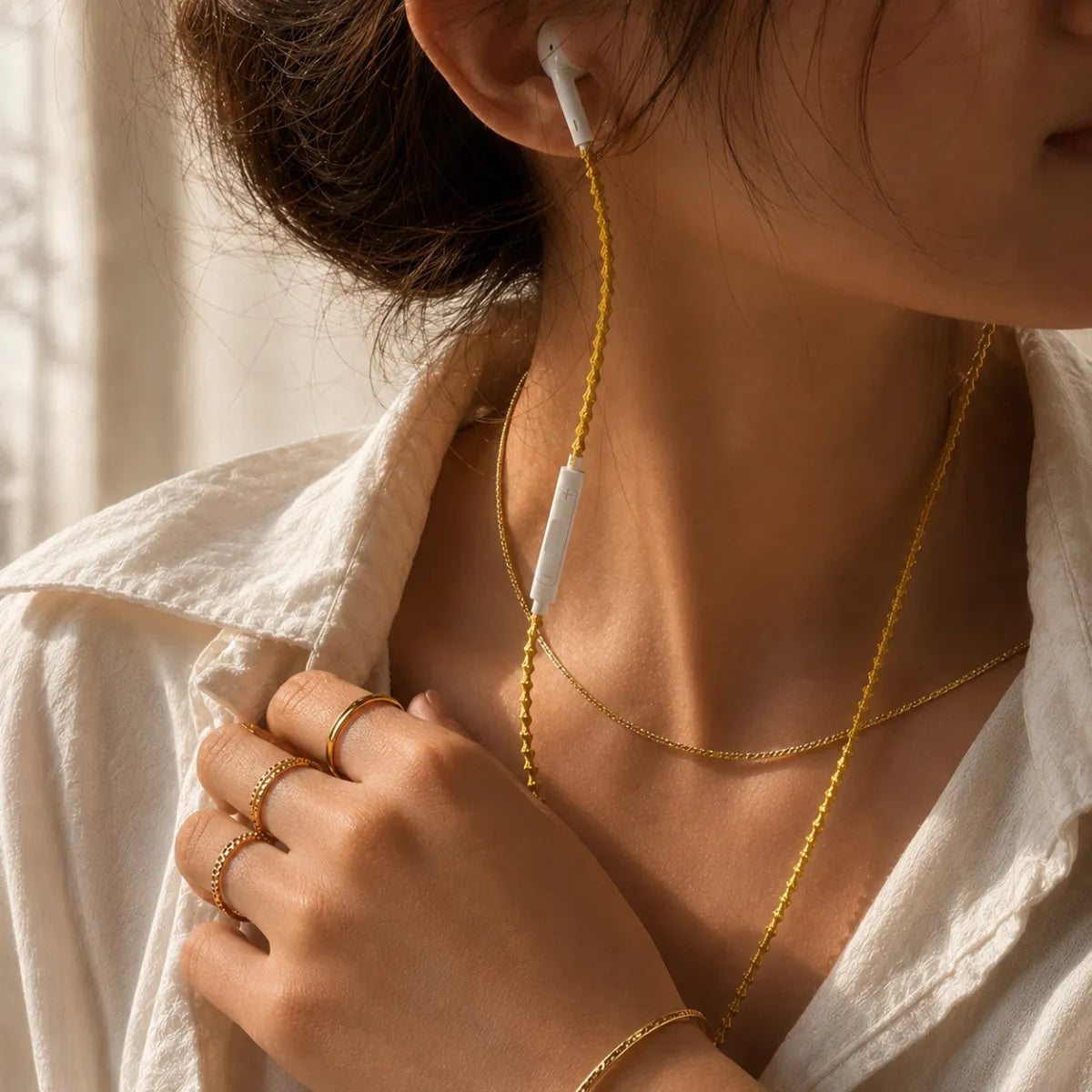  Layered gold jewelry detail with stacked gold rings, thin gold chain necklace, and gold wrapped earphones together at wrist and collarbone for all-gold layering aesthetic 