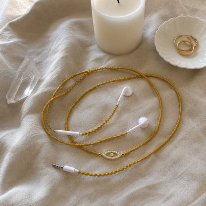  Gold evil eye earphones loosely coiled on washed beige linen beside quartz crystal, white candle and gold hoop earrings 