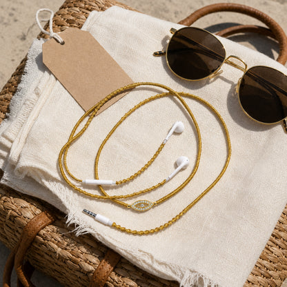  Top-down travel snapshot of gold evil eye earphones coiled on linen scarf and woven straw bag with gold sunglasses 
