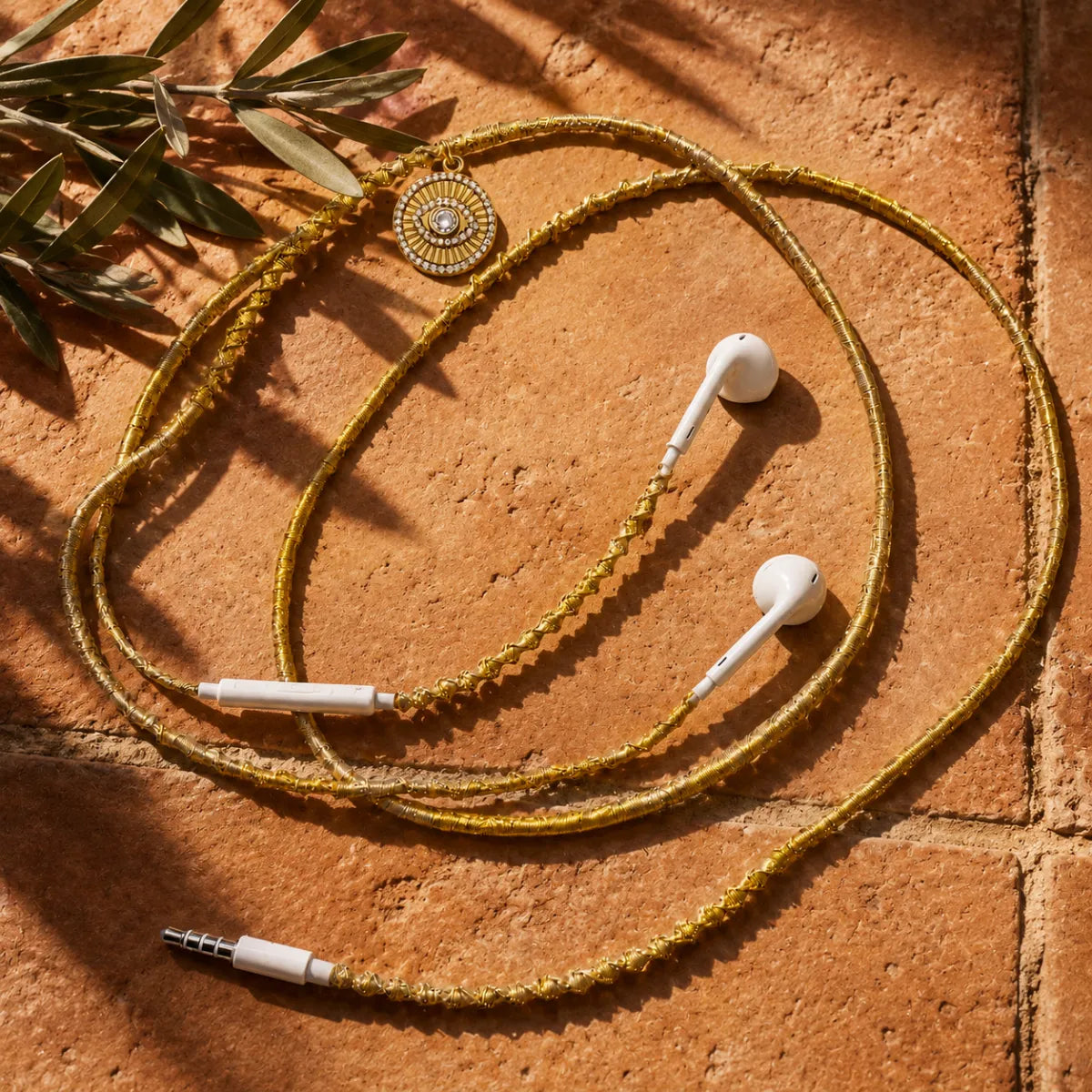  Gold evil-eye-medallion earphones on sun-warmed terracotta tile with soft-focus olive branch under strong Mediterranean sunlight, concept ad 
