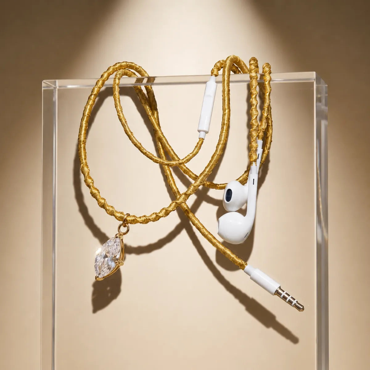  Gold wrapped earphones with marquise-cut crystal pendant draped over acrylic display block on warm cream gradient, fine-jewelry showcase ad 