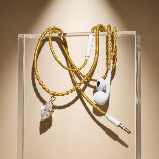  Gold wrapped earphones with marquise-cut crystal pendant draped over acrylic display block on warm cream gradient, fine-jewelry showcase ad 