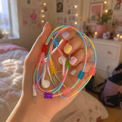 Hand holding a Gummy Bear Earphone Wrap with rainbow braided cord and colorful candy-inspired charms in a cozy bedroom.