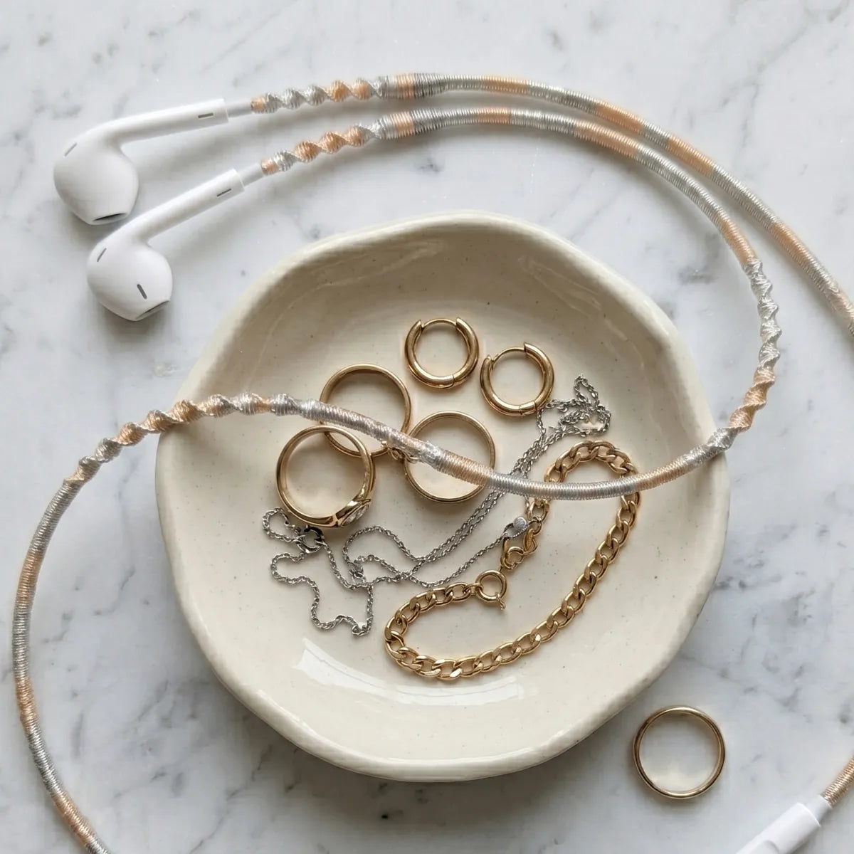  Cream ceramic jewelry tray with scattered gold and silver rings, hoop earrings, and chain necklaces with mixed metal wrapped earphones coiled through the assortment 