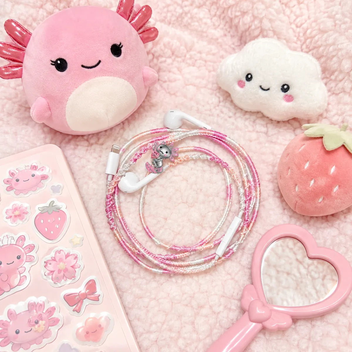  Kawaii pink axolotl earphone charm wrap surrounded by Squishmallow-style plushies and a pastel sticker book on a fluffy pink sherpa blanket 
