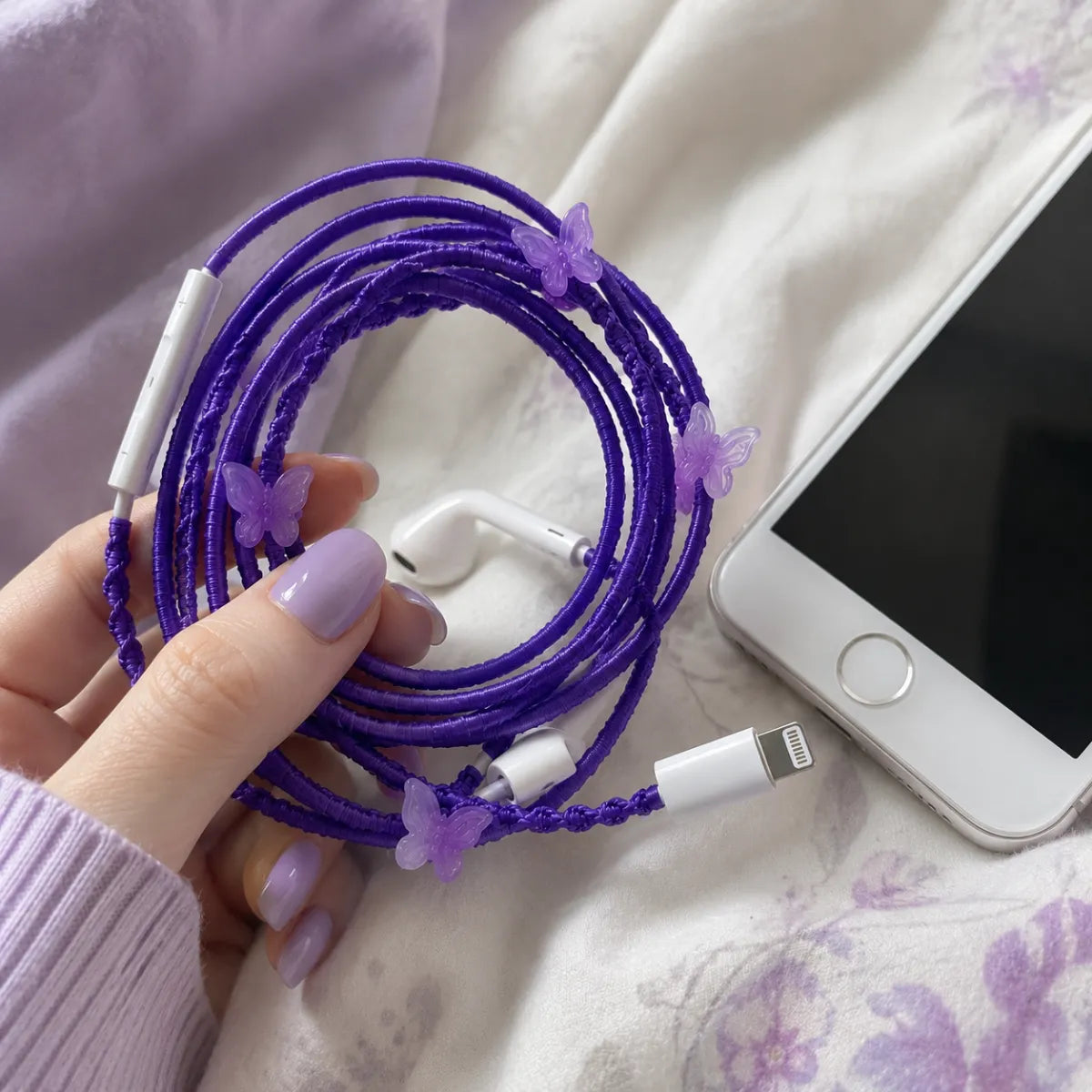  Hand plugging purple butterfly earphones into a white iPhone on a pastel bedsheet, glossy lavender nails, casual handheld lifestyle shot 