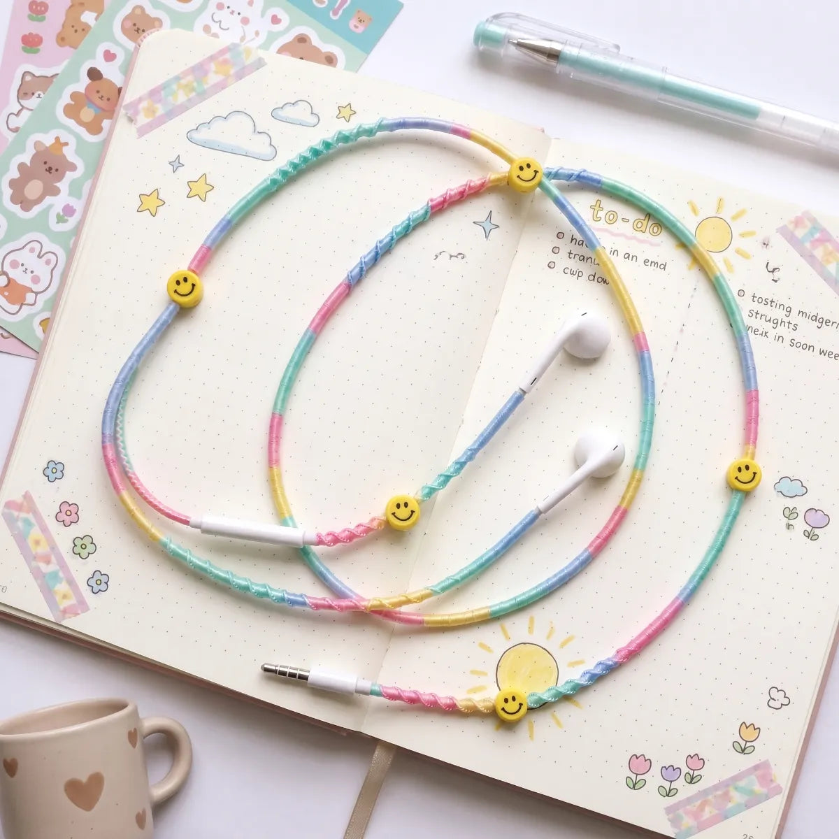  Pastel rainbow smiley earphones laid across an open bullet journal with washi tape, sticker sheets and a clear pen in soft daylight 