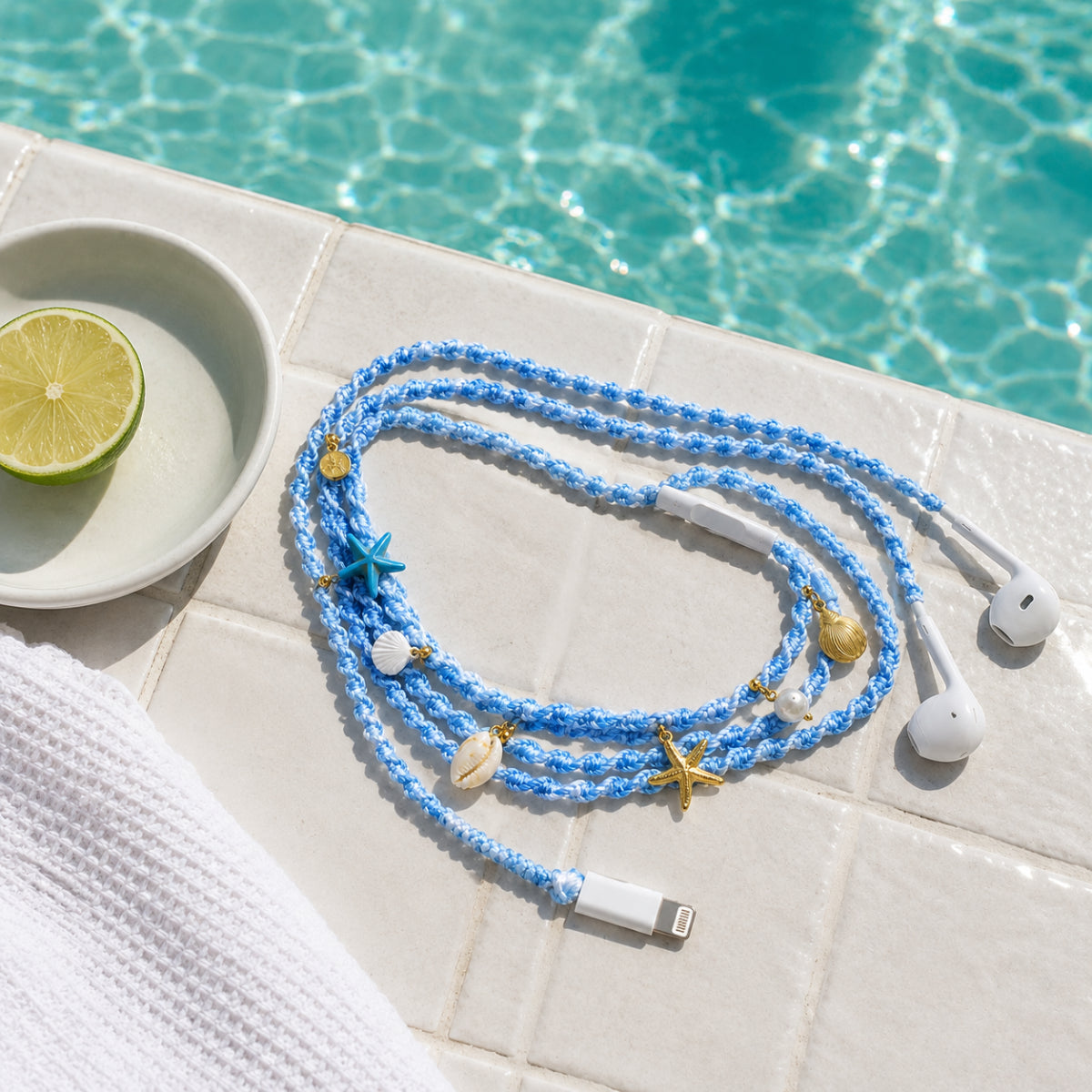  Blue and white braided shell earphones with gold charms lying on white poolside tiles beside turquoise pool water and lime slice 