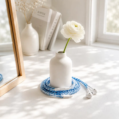  Sky blue crochet wrapped earphones coiled around the base of a small white vase with a single white blossom on a clean white desk in soft window light 