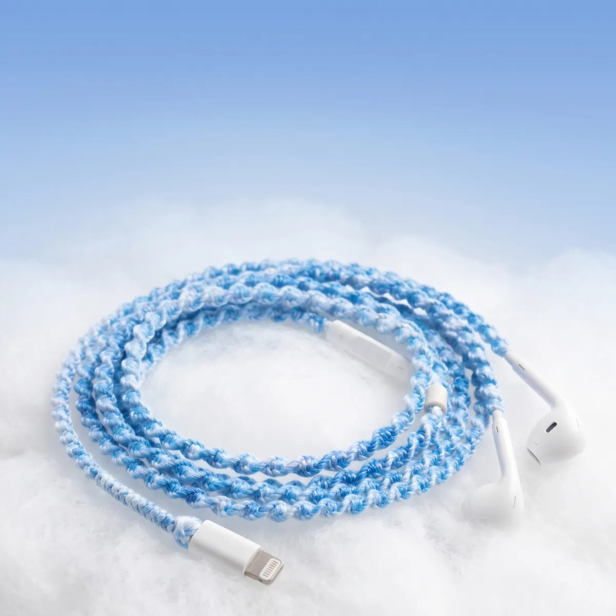  Sky blue crochet wrapped earphones with white Lightning connector resting on a soft white cloud-textured surface against a pale blue sky gradient for a piece of sky aesthetic ad 
