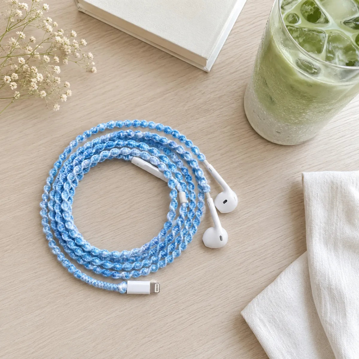  Top-down flat lay of sky blue crochet wrapped earphones beside a glass of iced matcha latte, a small white hardcover book, and a sprig of cream blossom on pale wood 