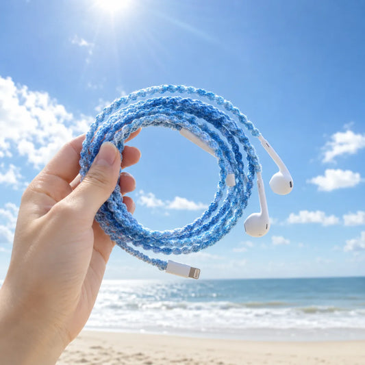 Hand holding sky blue crochet wrapped earphones with white Lightning connector against pale sand and clear sunny sky horizon for a soft summer coastal aesthetic 