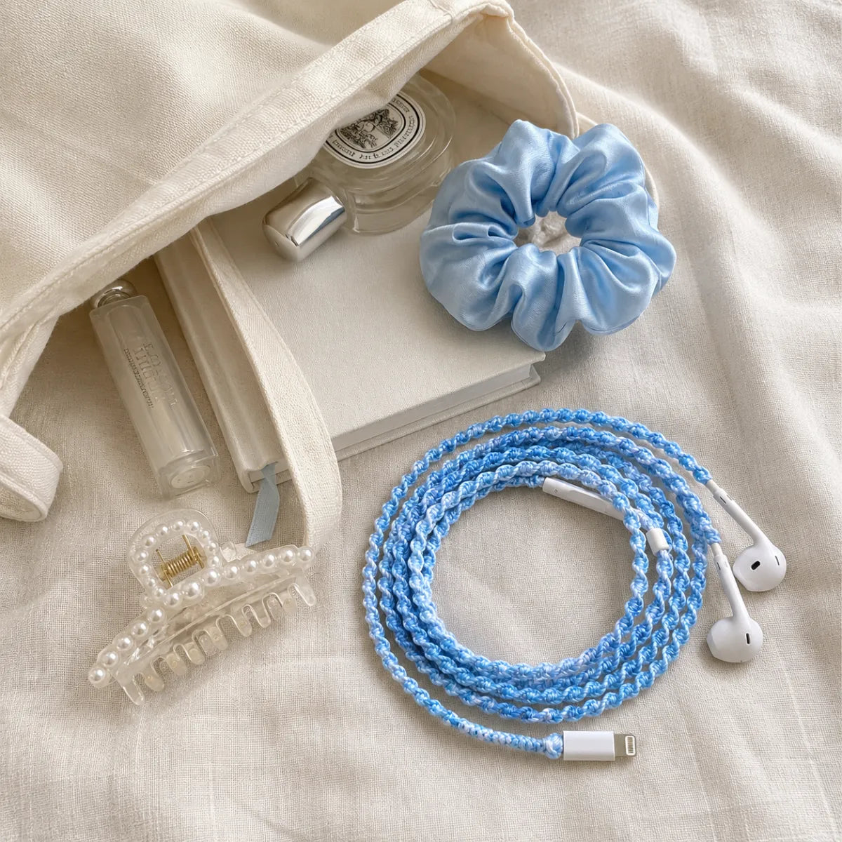  White canvas bag spilled flat lay with sky blue crochet wrapped earphones, white lip balm, pearl hair clip, small white notebook, and a sky blue silk scrunchie on cream linen 