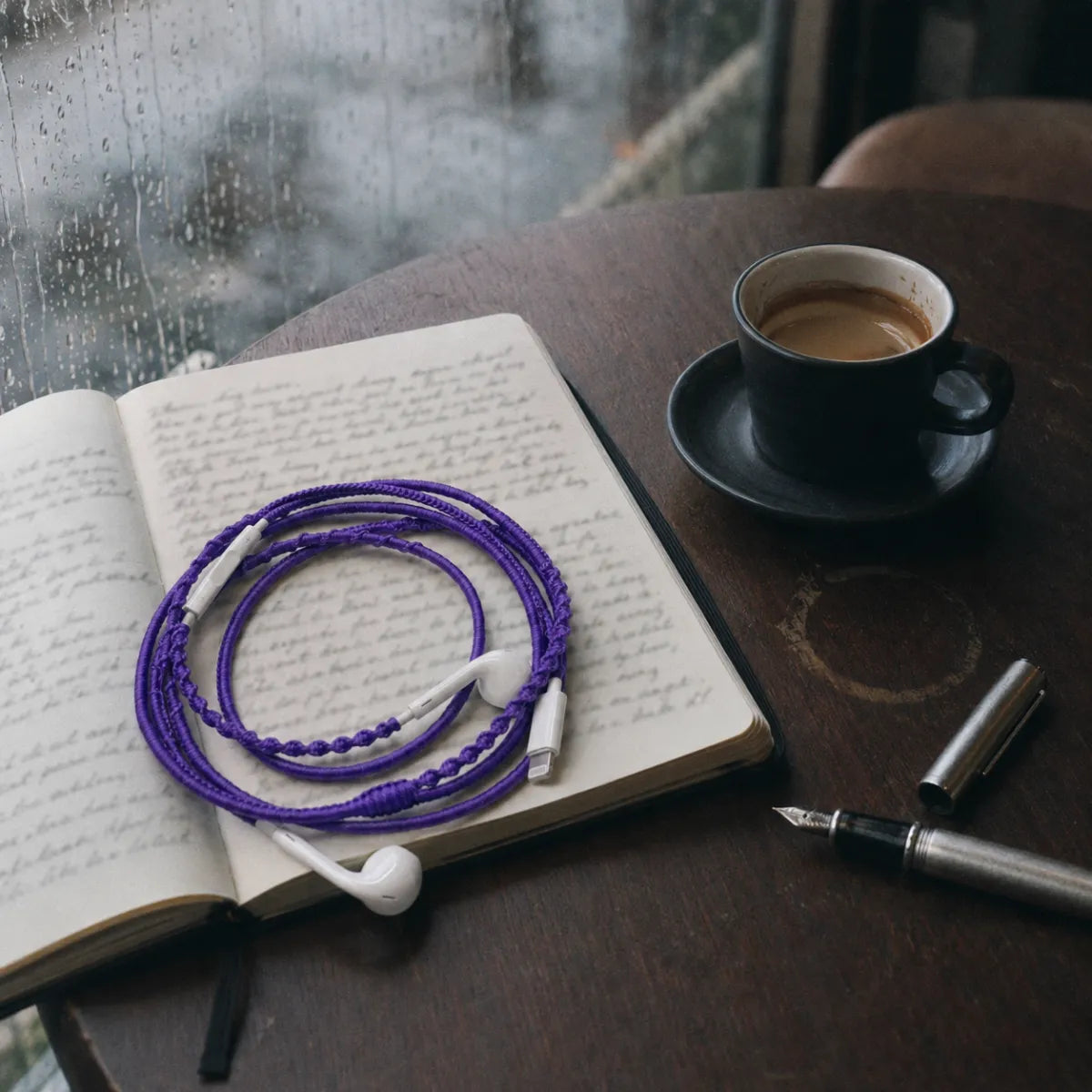  Violet earphone wrap resting on an open journal with handwritten notes beside an espresso cup and silver fountain pen at a rainy cafe 