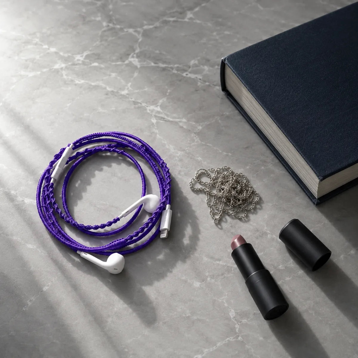  Deep violet earphone wrap coiled on cool gray marble with silver chain, dark cloth-bound book, and matte black lipstick in editorial quiet-luxury styling 