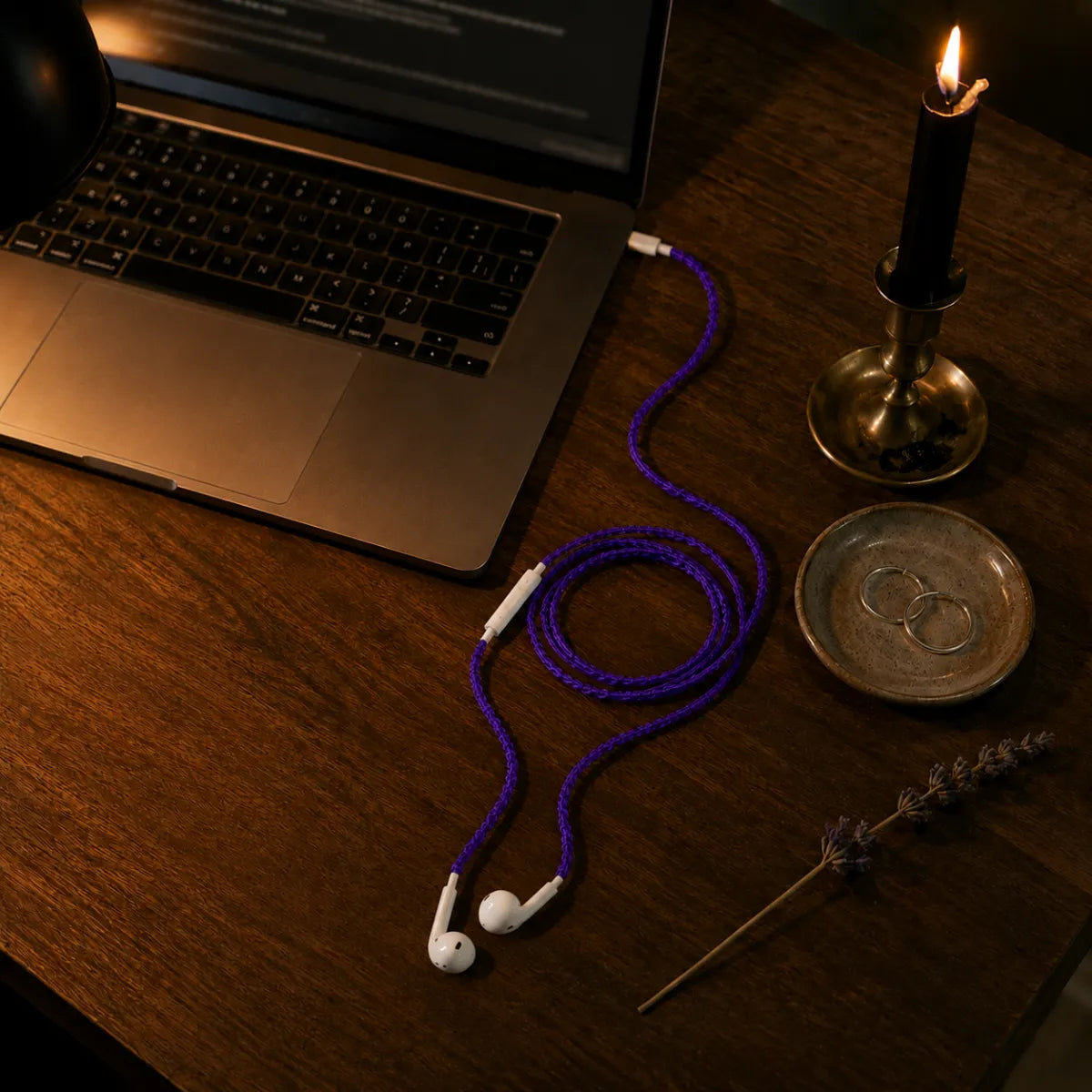  Violet earphone wrap connected to iPhone on a walnut desk with MacBook, silver earrings, candle, and dried lavender in warm lamplight 