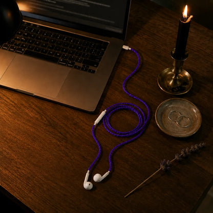  Violet earphone wrap connected to iPhone on a walnut desk with MacBook, silver earrings, candle, and dried lavender in warm lamplight 